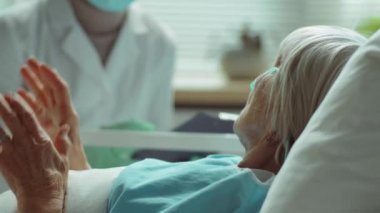 Yaşlı bir kadının hastane koğuşunda yatarken ve koruyucu maske takan kadın doktorla koronavirüs tedavisi görürken görüntüsünü kaldır.