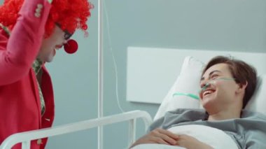 Kırmızı burunlu neşeli palyaço hastane koğuşunda terapi sırasında genç bayan hastayı güldürüyor.