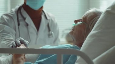 Korunaklı yüz maskesi takan erkek doktor, Coronavirus tedavisi sırasında onu desteklerken hastane koğuşunda yatağında uzanan yaşlı kadının ellerine dokunuyor.