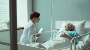 Laboratuvar önlüklü profesyonel bir kadın doktor elinde pano ve hastane koğuşunda yatarken yaşlı bir kadına tedavi planını açıklıyor.
