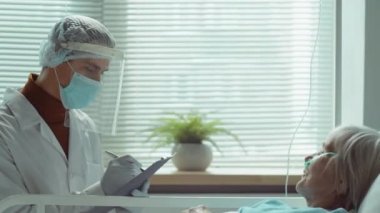 Koruyucu yüz kalkanı, maske ve eldiven giyen erkek doktor panoya bazı notlar yazıyor ve hastanede koronavirüs tedavisi gören yaşlı kadınla konuşuyor.
