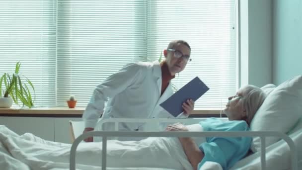 男医生带着剪贴板走进医院病房 坐在床边 与患有鼻腔疾病的老年妇女交谈 — 图库视频影像