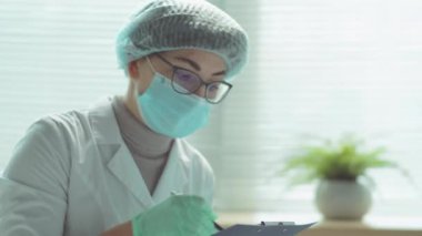Koruyucu maskeli kadın doktor, steril şapka ve eldivenli yaşlı bir kadınla konuşurken hastane koğuşunda oturuyor ve koronavirüs salgınında yardım ediyor.