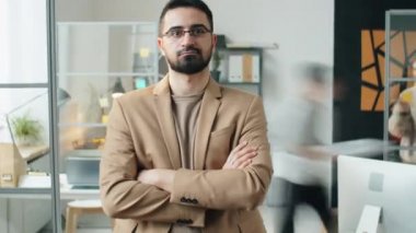 Gözlüklü ve bej takım elbiseli kendine güvenen işadamının iş günü açık ofiste dururken kolları çaprazlanmış bir şekilde kameraya poz verişi.