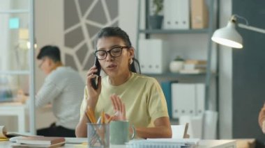 İspanyol iş kadınının iş günü açık ofiste masasında otururken cep telefonuyla konuşmasının görüntüsü.