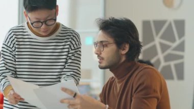 Genç Asyalı kadın ve Orta Doğulu erkek ofiste birlikte çalışırken iş belgelerini ve projeleri tartışıyorlar.