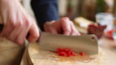 Erkek aşçının önlüklü elleri, ahşap kesme tahtasında kırmızı dolma biber doğrarken mutfak masasında yemek hazırlarken yakın plan çekimleri.