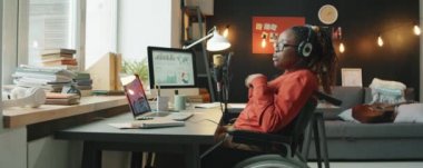 Özürlü genç Afrikalı bir kadın tekerlekli sandalyede dizüstü bilgisayarı ve mikrofonuyla oturuyor ve ev stüdyosunda iş podcast 'ini kaydederken konuşuyor.