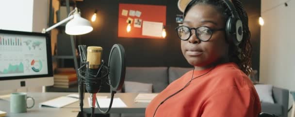 非洲裔美国女播音员头戴耳机坐在办公桌前 手持话筒 在家庭录音室摆姿势拍照的画像 — 图库视频影像