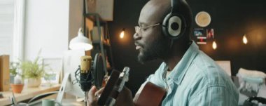 Ev stüdyosunda şarkı söylerken gitar çalıp mikrofonla şarkı söyleyen kulaklıklı Afrikalı Amerikalı bir müzisyen.