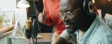 Ev stüdyosunda şarkı söylerken yanında dans eden heyecanlı kız arkadaşıyla birlikte gitar çalıp şarkı söyleyen kulaklı Afrikalı Amerikalı bir adam.