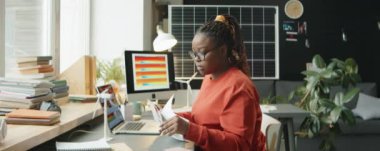 Afrika kökenli Amerikalı kadın mühendis ofis masasında sürdürülebilir enerji projesi üzerinde çalışırken bilgisayarında sınav kağıtları ve yazı yazıyor.