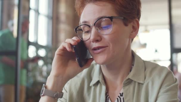 オフィスで働く傍ら 笑顔で携帯電話で話す眼鏡の経営者の女性をリヘッド — ストック動画