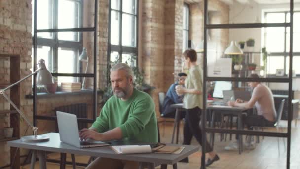 中世のビジネスマン灰色の髪とひげラップトップで作業し 現代のオープンスペースオフィスのデスクでクリップボードにメモを取る — ストック動画