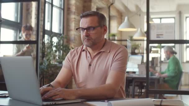 オフィスのデスクに座って 仕事中にノートパソコンを使用して眼鏡や損耗品で成熟したビジネスマン — ストック動画