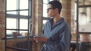 Güzel orta yaşlı iş kadını açık uzay ofisinde akıllı telefon kullanarak yürüyor, sonra masada oturuyor ve dizüstü bilgisayarla çalışıyor.