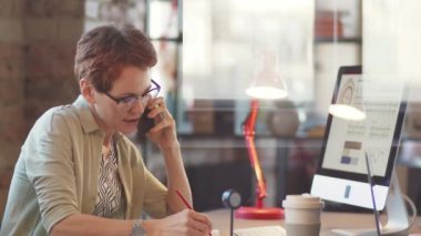 Cep telefonuyla konuşan ve masa başında çalışırken konuşma sırasında not alan iş kadınlarının cam ofis duvarından bak