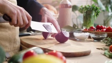 Erkek lokanta şefinin kırmızı soğan ampulünü iki parçaya ayırıp tahtadan yapılmış kasap bıçağıyla doğramaya hazırlandığı yakın plan çekimleri.