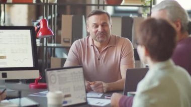 Orta yaşlı işadamı bilgisayar ekranındaki çizelgelere işaret ediyor ve ofiste iş arkadaşlarıyla tartışıyor.
