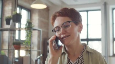 İş günü iş yerinde yürürken gülümseyen ve cep telefonuyla konuşan bir iş kadını.