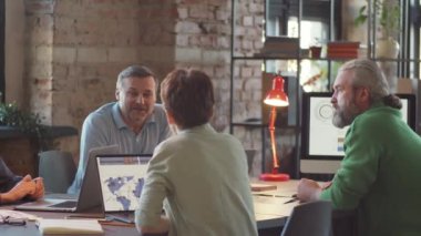 Orta yaşlı meslektaşlar ofiste toplantı masasında oturup iş projelerini tartışırlar.