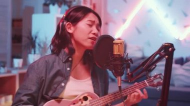 Evde renkli neon ışıklı bir şarkı kaydederken, kulaklıklı, gitar çalıp mikrofona şarkı söyleyen Asyalı genç bir bayan müzisyenin seçici odak noktası.