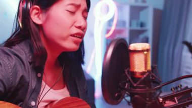 Asya kökenli genç bayan müzisyenin kulaklıkla gitar çalarken ve yoğunlaştırılmış mikrofonla şarkı söylerken ev stüdyoda renkli neon ışıklı bir şarkı kaydederken yakın plan çekimleri.