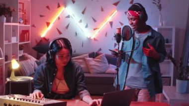 Çok ırklı müzik ikilisi ev stüdyosunda hip-hop kaydediyor: kulaklık takan havalı Afrikalı Amerikalı adam mikrofonda rap okurken Asyalı kadın DJ masasında karıştırma konsolu kullanıyor