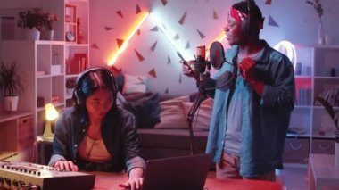 Genç arkadaşlar stüdyoda hip-hop kaydediyorlar: Asya 'lı bayan müzisyen masa başında karıştırma konsolu ve dizüstü bilgisayar kullanırken, kulaklıklı Afro-Amerikalı adam mikrofonda rap okuyor.