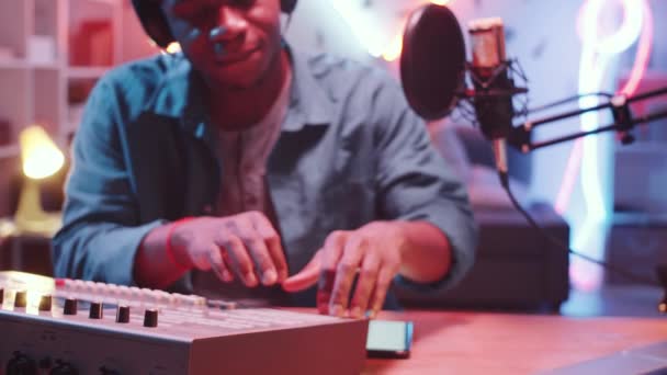 放大了年轻的非洲裔美国男性Dj坐在办公桌前的耳机中的镜头 他的头随着节奏移动 并在家里的录音室里用时髦的霓虹灯录制音乐时使用了混合控制台 — 图库视频影像