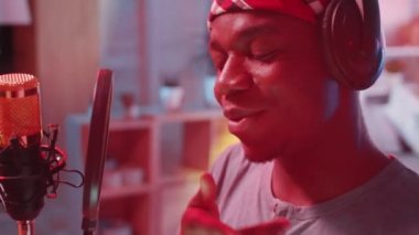 Genç Afrikalı Amerikalı rapçi, kulaklıkla stüdyoda renkli neon ışıklarıyla dolu bir mikrofon önünde hip-hop şarkısı kaydediyor.