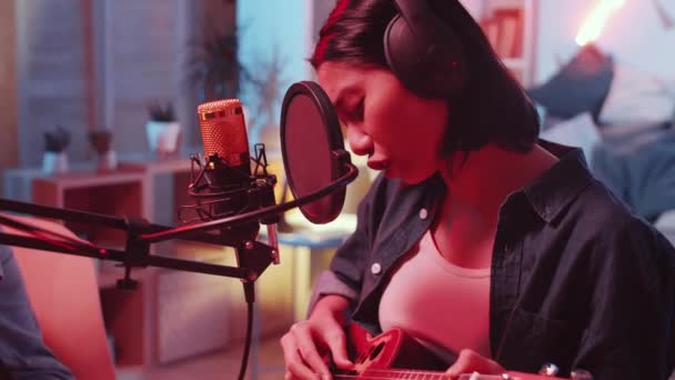 マイクでギターと歌を演奏するヘッドフォンで才能のあるアジアの女性の撮影を追跡しながら アフリカ系アメリカ人男性はネオンライトと自宅のレコーディングスタジオでデスクでミキシングコンソールを使用して — ストック動画