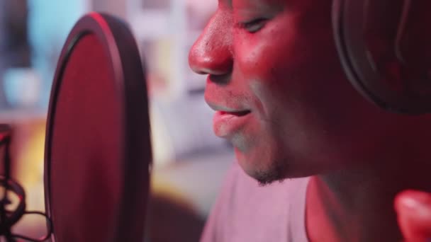 マイクの前でラップをしながら カラフルなネオンでいっぱいのホームスタジオでヒップホップの歌を録音している若いアフリカ系アメリカ人の男のクローズアップビュー — ストック動画