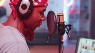 Afrika kökenli Amerikalı erkek hip-hop şarkıcısının mikrofonla rap yaparken ev stüdyosunda renkli neon ışıklarıyla müzik kaydederken yakın plan çekimleri.