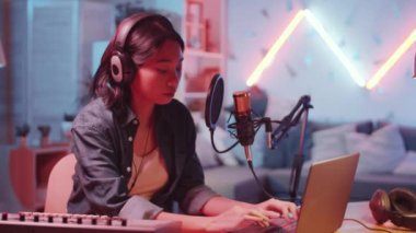 Kulaklıklı genç, güzel bir Asyalı kadın kameraya bakıyor ve masasında oturmuş neon ışıkla kayıt stüdyosunda mikrofon ve laptopu karıştırırken gülümsüyor.