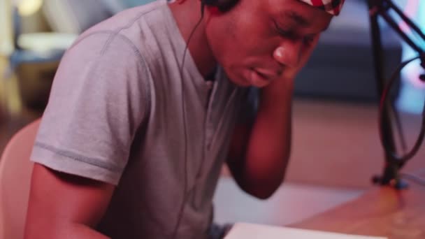 自宅のレコーディングスタジオのデスクに座っているヘッドフォンで若いアフリカ系アメリカ人のヒップホッププロデューサーのショットを閉じます 彼の頭を音楽に移動し ノートパッドで歌詞を書きます — ストック動画