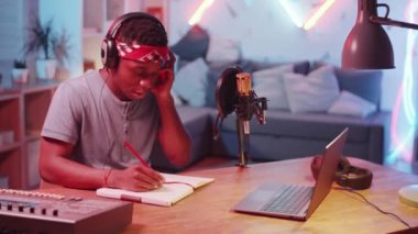 Kulaklıklı genç Afrikalı Amerikalı bir adam kayıt stüdyosunda masasında oturuyor, başını müziğe çeviriyor ve yeni bir şarkı üzerinde çalışırken şarkı sözü yazıyor.