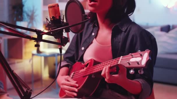 アジアの女性歌手がギターを弾き マイクに歌を歌いながら カラフルなネオンでホームスタジオの友人の助けを借りて曲を録音するシーンをクローズアップ — ストック動画