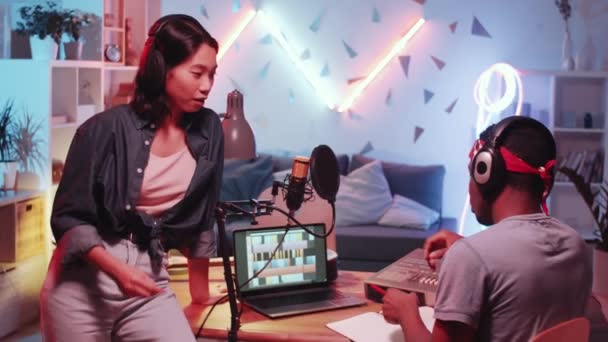 若いアフリカ系アメリカ人の男性ソングライターがデスクに座り アジア系女性ミュージシャンと音楽について話し合いながら カラフルなネオンの光でいっぱいのホームレコーディングスタジオで新しい曲に取り組みます — ストック動画