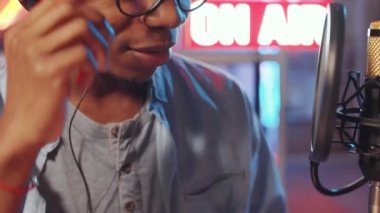 Siyah erkek radyo sunucusunun mikrofona gülümseyip konuştuğu yakın plan çekimleri. Stüdyoda arka planda ışık ve ışık var.