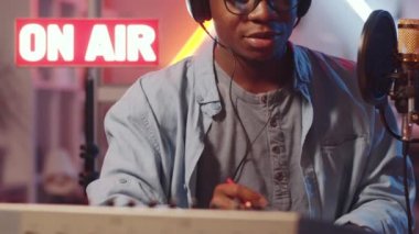 Afrika kökenli Amerikalı bir erkek sunucunun radyo stüdyosunda neon ışığı ve ışıklı ışıklarla mikrofona konuşurken orta bölüm görüntüsü.