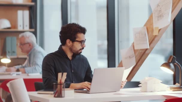 中東民族の若い男性建築家は オープンスペースオフィスの机に座って ノートパソコンを使用して 作業中に文書をチェックします — ストック動画