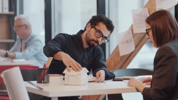 オフィスのテーブルに座っている若い中東の男性建築家と彼の白人女性の同僚は プロジェクトに取り組んでいる間 家のモデルのデザインを笑顔で話し合います — ストック動画