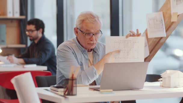 ガラスのシニア男性建築家とオープンスペースのオフィスでデスクで作業しながら ラップトップ上のオンラインビデオ通話を介して建設計画を議論カジュアルウェア — ストック動画