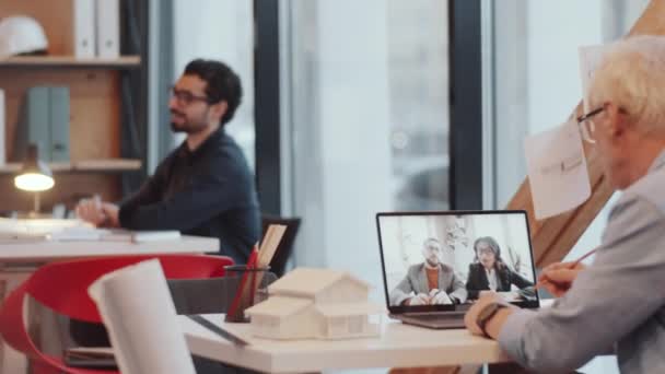 オープンスペースオフィスのデスクに座ってノートを取り ラップトップ上のオンラインビデオ通話を介して2人の同僚と議論を持つシニア男性建築家 — ストック動画