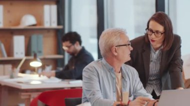 Genç bayan mimar ofis masasına yaslanıp dizüstü bilgisayardaki projeyi yaşlı bir erkek meslektaşla tartışıyor ve sonra karışık yaştaki iş arkadaşları kameraya bakıp gülümsüyor.