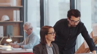 Orta Doğulu genç bir erkek mimar ofisteki neşeli beyaz kadın meslektaşına doğru yürüyor, masaya yaslanıyor ve dizüstü bilgisayarında projeyi tartışıyor.