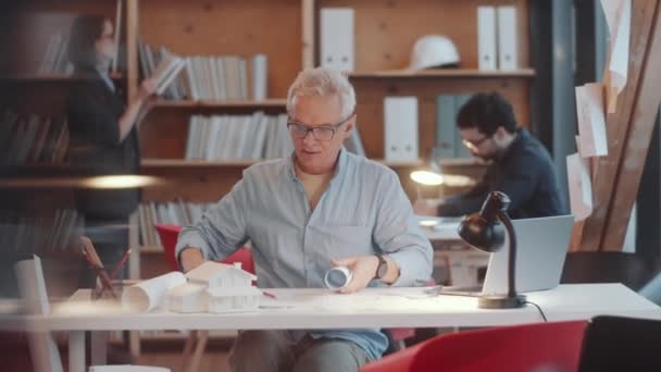 Arquiteto Masculino Sênior Óculos Casualwear Andando Escritório Sentado Mesa Desenrolando — Vídeo de Stock
