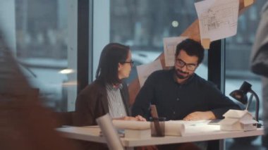 Kafkas kadın mimar ve ortadoğulu erkek iş arkadaşı birlikte masa başında oturmuş inşaat planını tartışıyorlar ve modern ofiste birlikte çalışıyorlar, yerden tavana geniş pencereleri var.