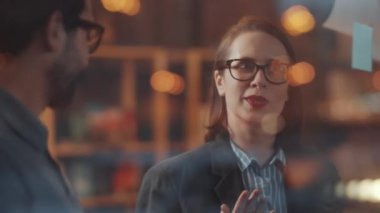 Ofisteki cam duvara yapıştırıcı notlar koyarken orta doğulu erkek meslektaşlarıyla gülümseyen ve sohbet eden neşeli beyaz iş kadını.